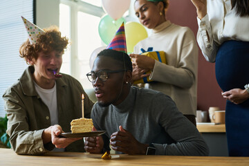 Young businessman blowing burning candle on piece of tasty birthday cake after making wish while...