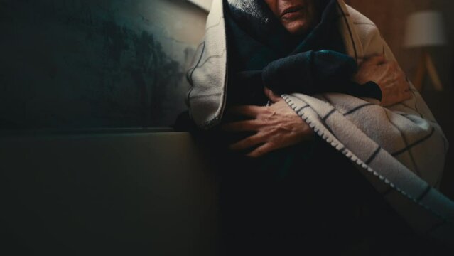 Poor old woman wrapped in a blanket warming up near electric heater in cold room