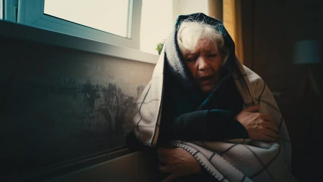 Frozen senior woman wrapped in blankets trying to warm up near electric heater
