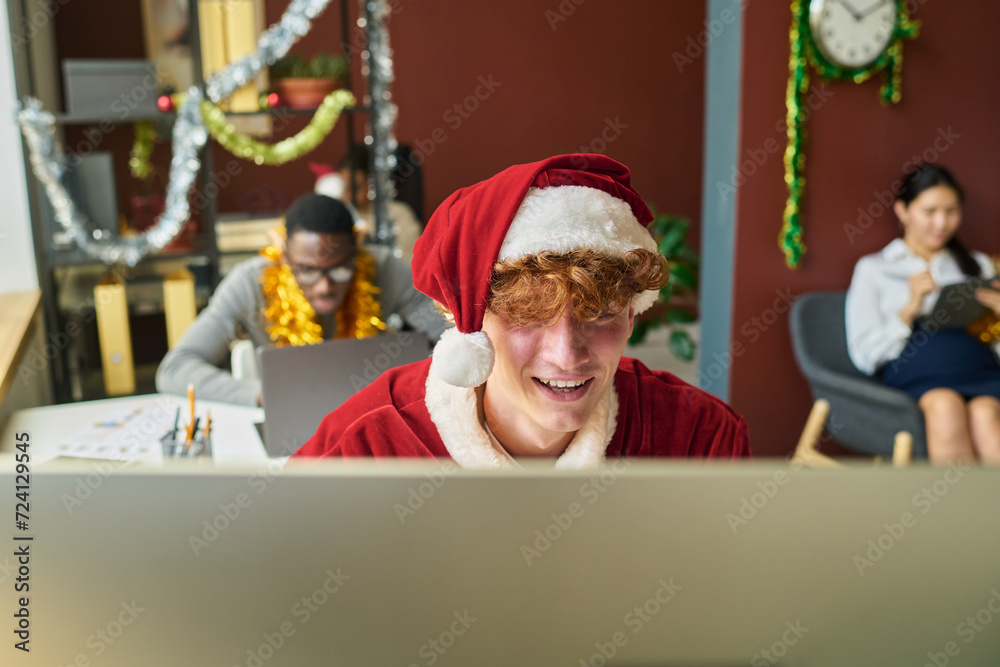 Poster young male office worker in costume of santa claus sitting in front of screen of desktop computer an