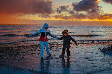 Mother and son in winter suits standing on slippery ice by lake shore at sunset