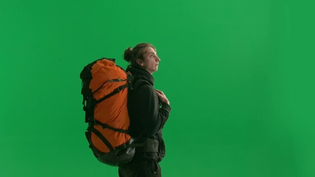 Side view of a male traveler with a large hiking backpack on his back close up. The man is standing, looking around and pointing his index finger to the side and down. Hiker in studio on green screen.