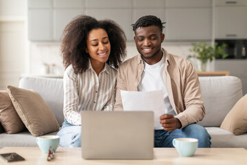 Happy black spouses paying bills via laptop holding papers indoor