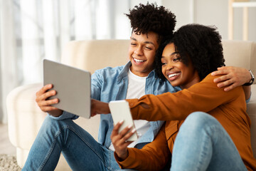 Smiling young black couple winners, with smartphone, tablet, sit on floor