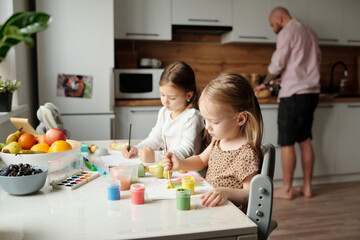 Adorable little girls sitting by kitchen table and painting with gouache and watercolors while their father preparing breakfast for family - obrazy, fototapety, plakaty