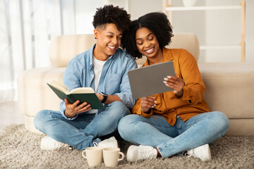 Fototapeta premium Seated comfortably on the floor, a young woman with a tablet and a man with a book share a moment