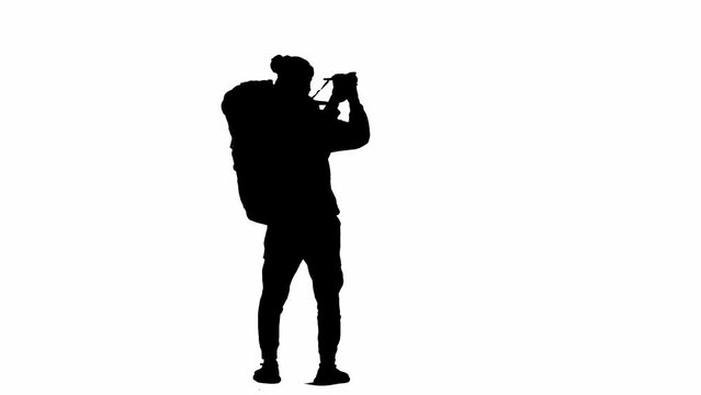 Black silhouette of a male traveler taking photos on a camera during a hike. A male traveler stands on white isolated background. Concept of travel, active rest, hiking. Rear view.