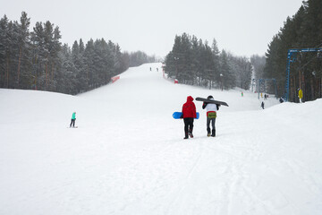 Panoramic view of a ski slope in snowfall. In the foreground are two snowboarders going up the slope, with a T-bar on the right. Copy space.