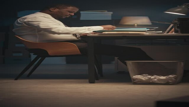Vertical shot of African American male entrepreneur making mistake in document filling, crumpling sheet od paper and throwing it into office bin beside his desk, working late at night