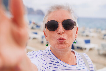 A senior woman with grey hair, in sunglasses taking a selfie on a beach 