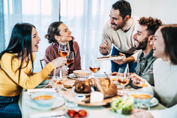 Happy group of friends having dinner party at home - Cheerful young people having lunch break together - Life style concept with guys and girls celebrating thanksgiving - Man serves vegetables - obrazy, fototapety, plakaty