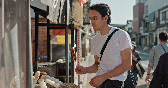 Japanese man, food and street vendor with kebab on travel, walk or tourism for taste, culture and product. Person, customer and shop for dango snack on sidewalk, vacation and urban road in Japan