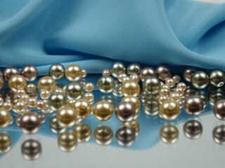 Pearl beads of various colors lie on the mirror and are reflected in it against the background of blue satin fabric.