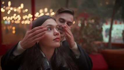 Loving young man coming up from behind and covering eyes of girlfriend on footpath. Romantic couple in trendy clothes embracing and kissing in illuminated city on Christmas night.