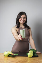 Smiling pretty athletic woman in sportswear holding glass of healthy green vegan smoothie. banana, spinach, apple and avocado on a table