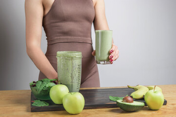 Unrecognizable athletic woman in sportswear holding glass of healthy green vegan smoothie. banana, spinach, apple and avocado on a table
