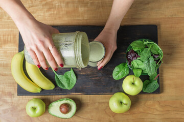 Woman's hands pouring healthy green vegan smoothie into a glass on a table with banana, spinach,...