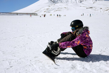 Fototapeta na wymiar Girl or boy in ski helmet, sunscreen mask and balaclava sits on snow and fix the snowboard bindings against the backdrop of snow-covered mountain ski slope and a cloudy sky. Winter. Sport and travel 