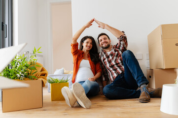 Beautiful young couple expecting a baby just moved into an empty apartment, sitting among cardboard...