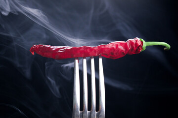 Red chili pepper on a fork with smoke on a black background.