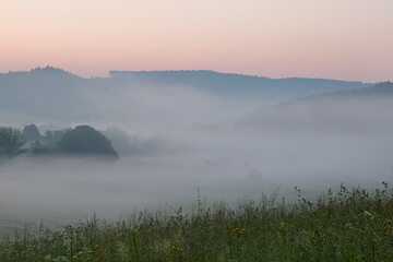Morning fog in the countryside, before sunrise. In the foreground is a meadow and in the background forests.