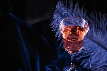 Glass of rose sparkling wine and large blue feather on dark blue velour background. Beautiful glamorous romantic greeting card