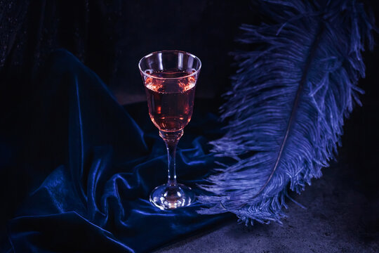 Glass of rose sparkling wine and large blue feather on dark blue velour background. Beautiful glamorous romantic greeting card