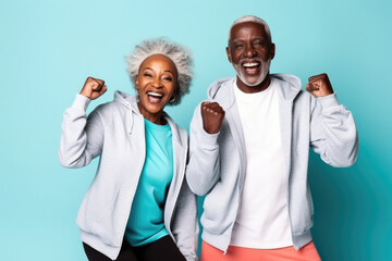 Portrait of Elderly afro-american smiling couple posing raising hands triumphantly over blue background. Mature adults wear activewear sportswear Sport fashion clothes for retired people man and woman