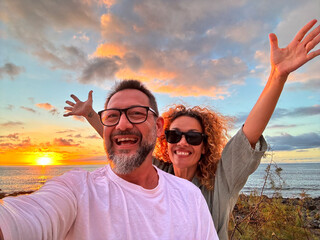 Crazy happy couple of tourist taking selfie picture at the beach with amazing sunset in background. Beach and ocean lifestyle people. Travel and destination. Outdoor leisure activity man and woman
