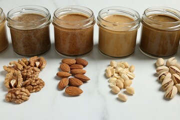 Tasty nut butters in jars and raw nuts on white marble table, closeup