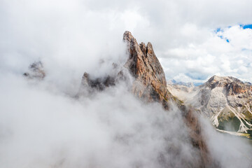 Dense fog descends onto Seceda mountain peaks on sunny summer day. Giant ridgeline with bare peak...