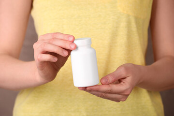 Woman holding blank white bottle with vitamin pills against light brown background, closeup