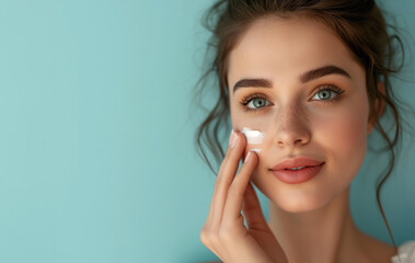 Smiling young woman applying facial cream. Close-up beauty portrait with skincare product on cheek, light blue background. Skincare and wellness concept. Design for banner with copy space or cosmetic.