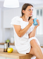 Fototapeta premium Portrait of relaxed young woman sitting on kitchen countertop, drinking coffee or tea in morning