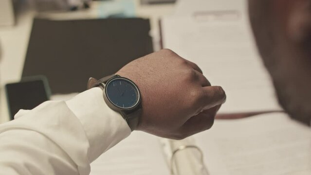 Over shoulder of African American businessman taking video of documents on desk and looking at wristwatch while working overtime in office at midnight