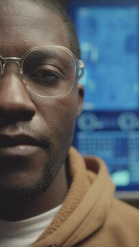 Vertical half face portrait of young Black male software engineer in eyeglasses looking at camera with computer data on blue screen in background
