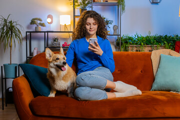 Happy young woman sitting on sofa using smartphone beside corgi dog in living room at home. Beautiful female girl text messaging and using social media via mobile phone during weekend in apartment.