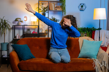 Excited young woman with curly hair having fun dancing and moving to rhythm sitting on sofa in...