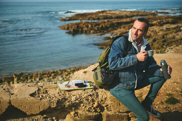 Happy adventurer sitting by Atlantic beach, smiles looking aside, holds a thermos, enjoying his coffee break outdoor