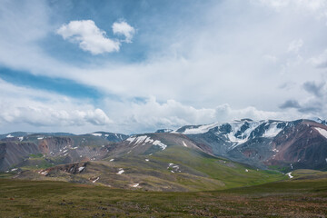 Dramatic alpine view from stony grassy hill to large snow-capped mountain range under cloudy gray...