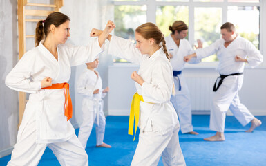 In gym, adolescence girl in kimono learns to fight and, paired with mother, practices striking. Family karate coach conducts classes for parents and children