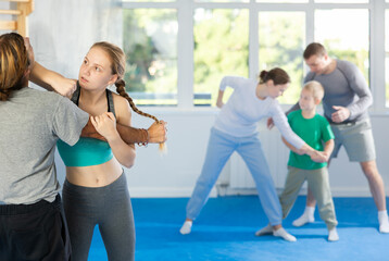 Fototapeta premium Man and teen girl at group self-defense lesson are learning new technique of blowing to chin, trainer is practicing technique with other pair in background