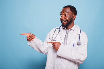 Portrait of cheerful male physician dressed in lab coat and stethoscope pointing next to him on isolated blue background. Smiling african american doctor gesturing with index finger to his right side.