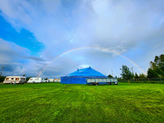 Fototapeta na wymiar Vibrant blue circus tent nestled under a magnificent double rainbow, set against a lush green field near a tranquil forest, with billowing white clouds and a clear blue sky