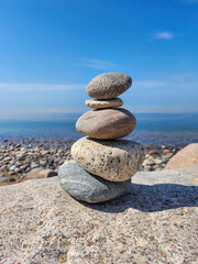 Tranquil Scene of a Stack of Smooth and Round Stones of Various Sizes Arranged on the Rocky Shore of a Calm Blue Lake, Set Against a Cloudless Sky Background in Bright Sunlight