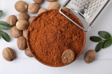 Nutmeg powder in bowl, seeds, grater and green branches on white table, flat lay