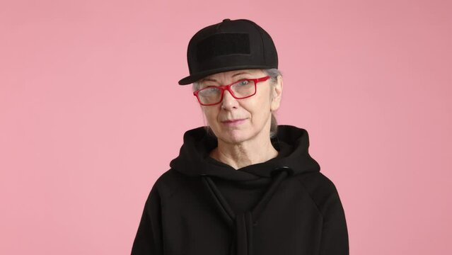 A mature woman in a black cap makes a stern cut-off gesture across her throat, indicating a strong refusal or the end of a matter, against a plain pink background. High quality 4k footage