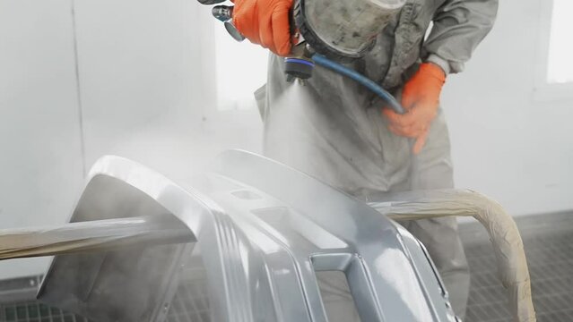 Modern car painting. Man is using spraying machine, holding it in hand, dressed in protective uniform. Master is painting part of automobile in car service.