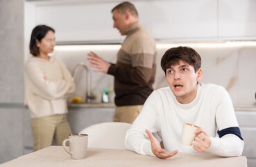 Obraz premium Young guy sad while sitting at table during family quarrel between adult man and young woman