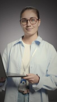 Vertical medium studio portrait of young Caucasian female programmer wearing eyeglasses and ID badge om her neck standing with open laptop on grey background and smiling at camera